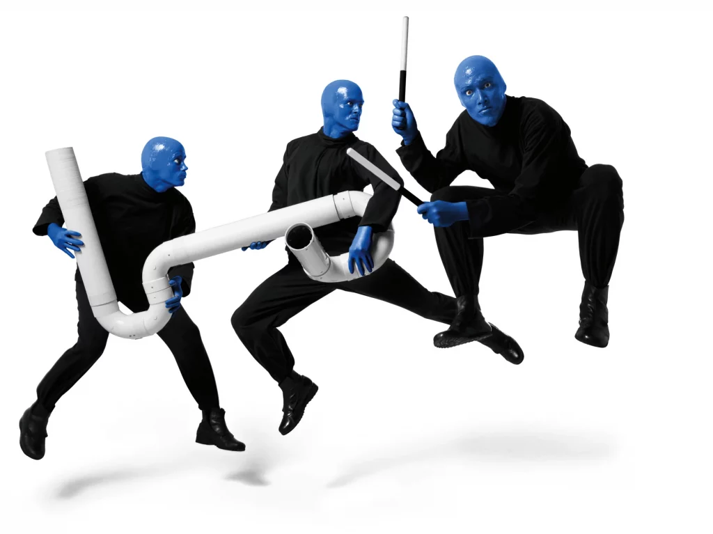 Blue Man Group Berlin - 3 Männer mit blauen Köpfen und ganz in schwarz gekleidet tanzen in der Luft vor weißem Hintergrund