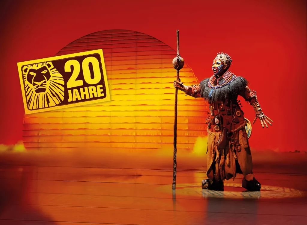 Disneys Der König der Löwen Hamburg - Rafiki mit Stab vor der aufgehenden Sonne und dem Logo 20 Jahre
